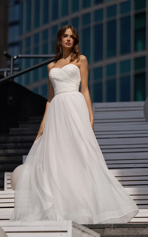 Robe de mariée en tulle à décolleté croisé et fronces, coupe trapèze, ornée de perles à la taille.