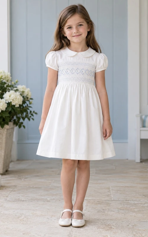 Robe de demoiselle d'honneur blanche simple et modeste, arrivant au genou, à col Claudine, manches bouffantes compactes, ornée de smocks bleus à losanges et boutonnée au dos.