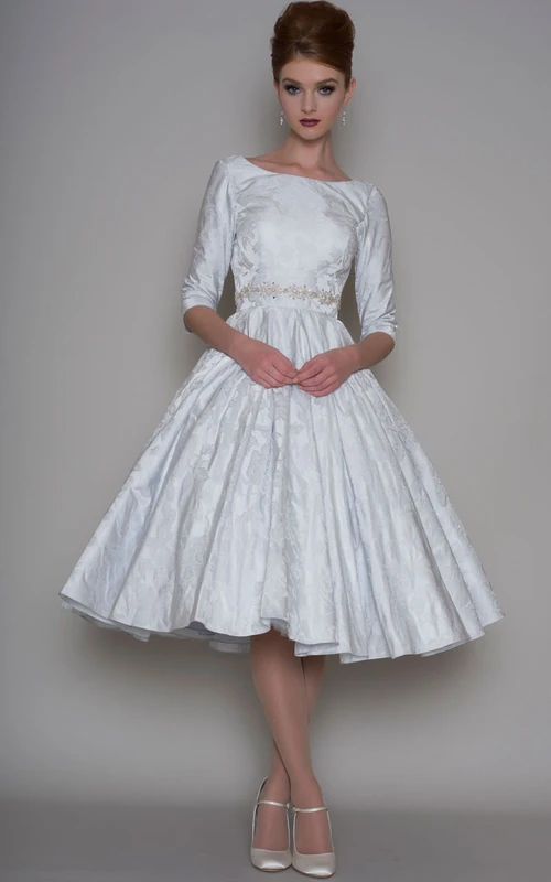 Vestido de novia de satén con escote barco, largo midi y corte en A, mangas tres o cuatro, adornado con joyas.