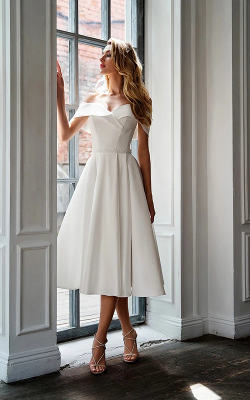 Vestido de novia de satén con escote Bardot, corte en A, largo hasta la rodilla y cintura adornada con cuentas.