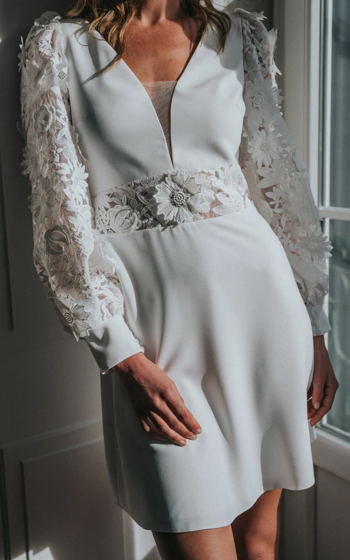Robe de mariée courte moderne à manches longues et col en V, ornée d'appliqués et d'une découpe en forme de goutte.