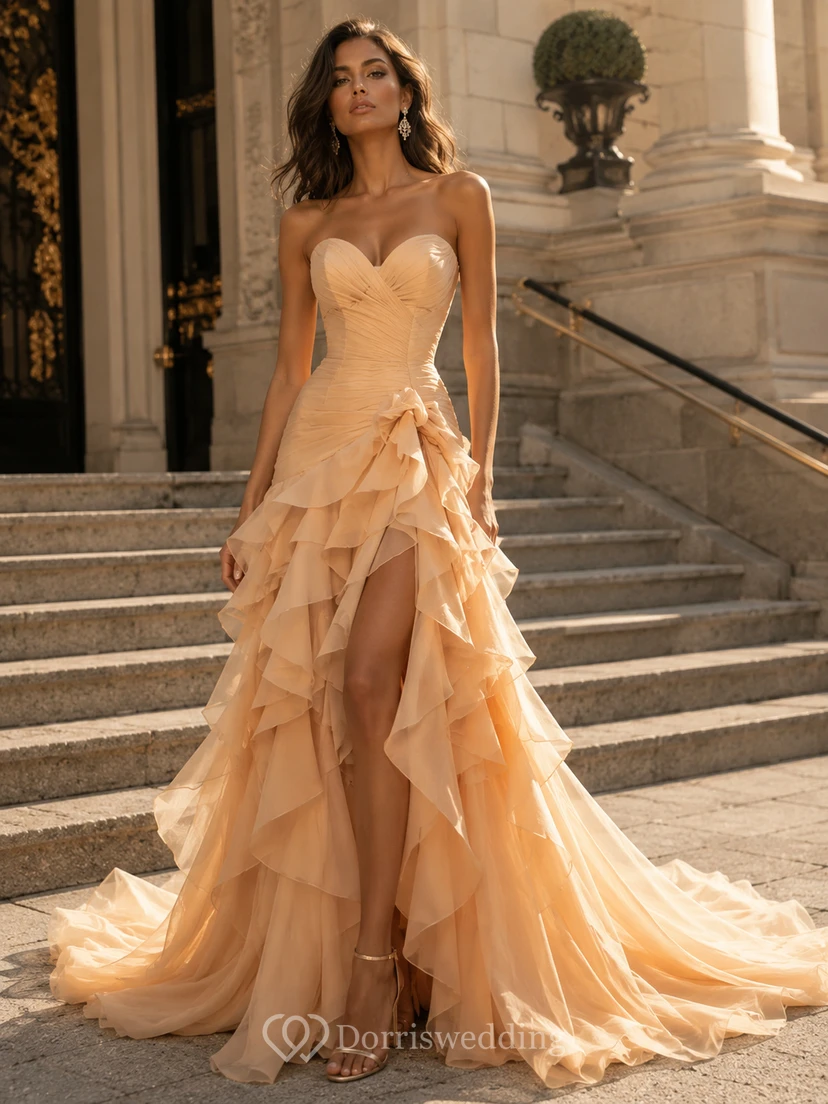 Vestido de fiesta de tul color champán, sin tirantes, con escote corazón, corte sirena, cola larga, volantes y capas.