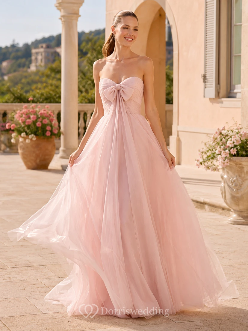 Robe de mariée en tulle rose poudré à fines bretelles, décolleté cœur et nœud oversize, coupe trapèze, bretelles spaghetti et bustier.